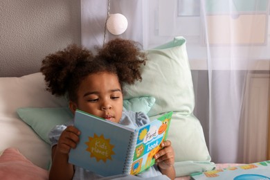 Photo of African American girl reading book on bed at home