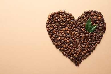 Photo of Heart shaped pile of coffee beans and fresh green leaves on light orange background, top view with space for text