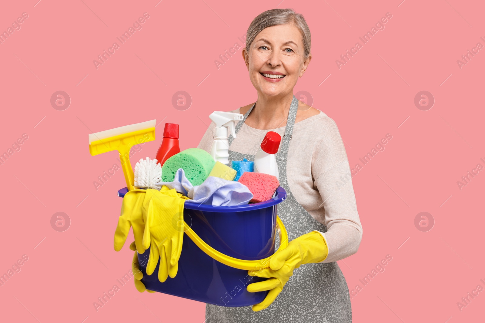 Photo of Happy housewife holding bucket with cleaning supplies on pink background