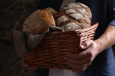 Photo of Man holding wicker basket with different types of bread on dark background, closeup