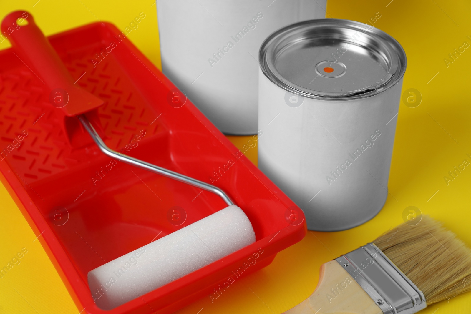 Photo of Cans of orange paint, brush, roller and container on yellow background, closeup