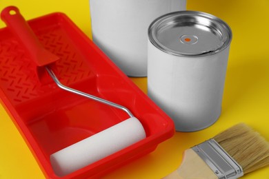 Photo of Cans of orange paint, brush, roller and container on yellow background, closeup