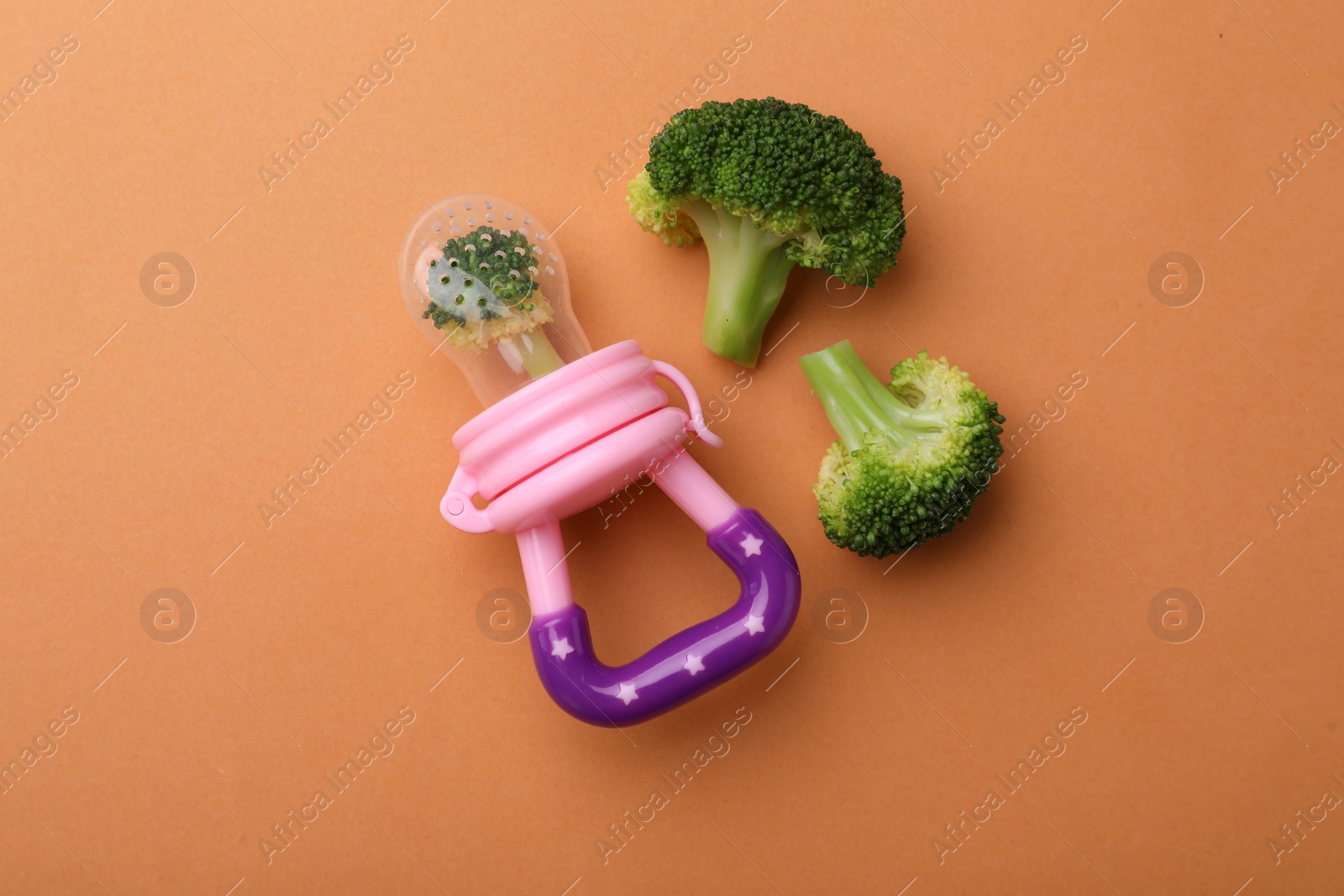 Photo of Nibbler with boiled broccoli on pale orange background, flat lay. Baby feeder