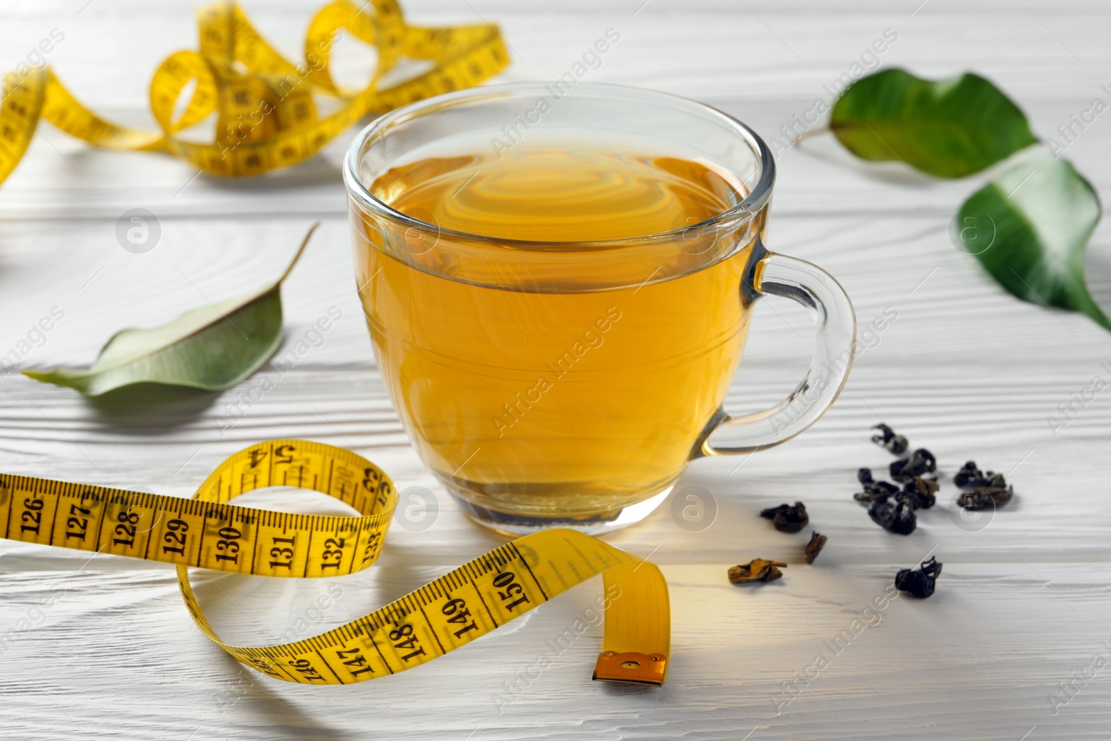 Photo of Cup of herbal diet tea, measuring tape and green leaves on white wooden table, closeup. Weight loss concept