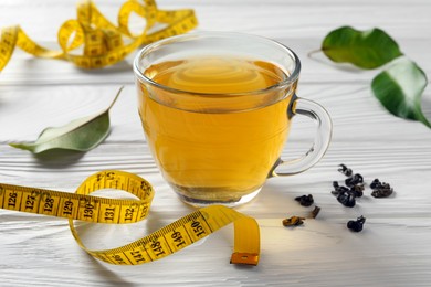 Photo of Cup of herbal diet tea, measuring tape and green leaves on white wooden table, closeup. Weight loss concept