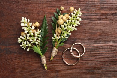 Small stylish boutonnieres and rings on wooden table, flat lay