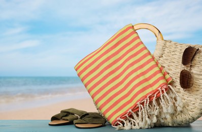 Beach bag with towel, slippers and sunglasses on light blue wooden surface near seashore