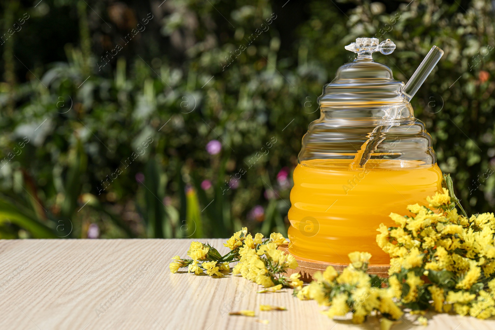 Photo of Delicious fresh honey and beautiful flowers on wooden table in garden, space for text