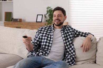 Photo of Happy man changing TV channels with remote control on sofa at home
