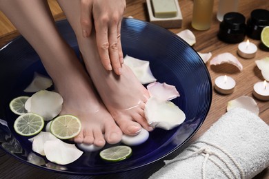 Photo of Woman soaking her feet in bowl with water, petals and lime slices on wooden floor, closeup. Spa treatment