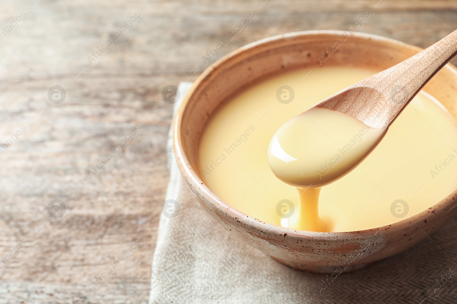 Photo of Spoon of pouring condensed milk over bowl on table, closeup with space for text. Dairy products