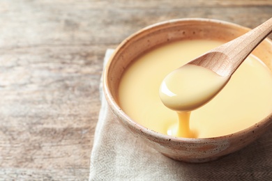 Photo of Spoon of pouring condensed milk over bowl on table, closeup with space for text. Dairy products