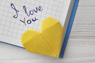 Photo of Notebook with written words I LOVE YOU and origami heart on white wooden table, top view