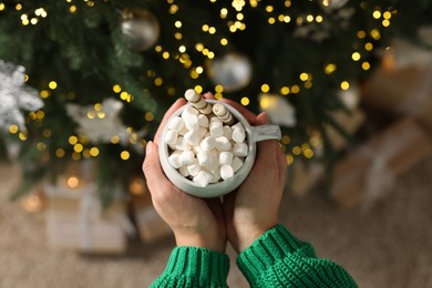 Photo of Woman holding cup of delicious Christmas cocoa with marshmallows and wafer sticks near fir tree, top view