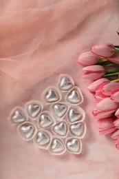 Heart made with delicious chocolate candies and beautiful tulips on pink table, flat lay