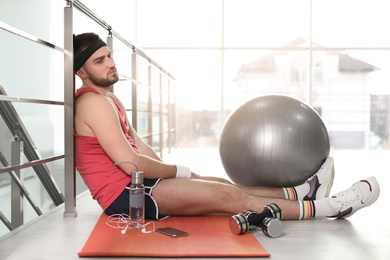 Photo of Lazy young man with sport equipment on yoga mat indoors