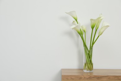 Beautiful calla lily flowers in glass vase on wooden table near white wall. Space for text