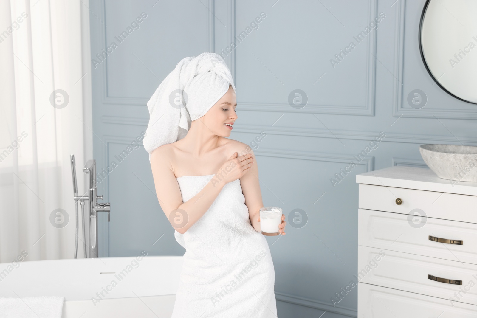 Photo of Beautiful young woman applying body cream onto shoulder in bathroom