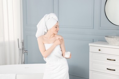 Beautiful young woman applying body cream onto shoulder in bathroom