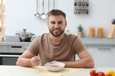 Young man eating tasty vegetable soup at table in kitchen