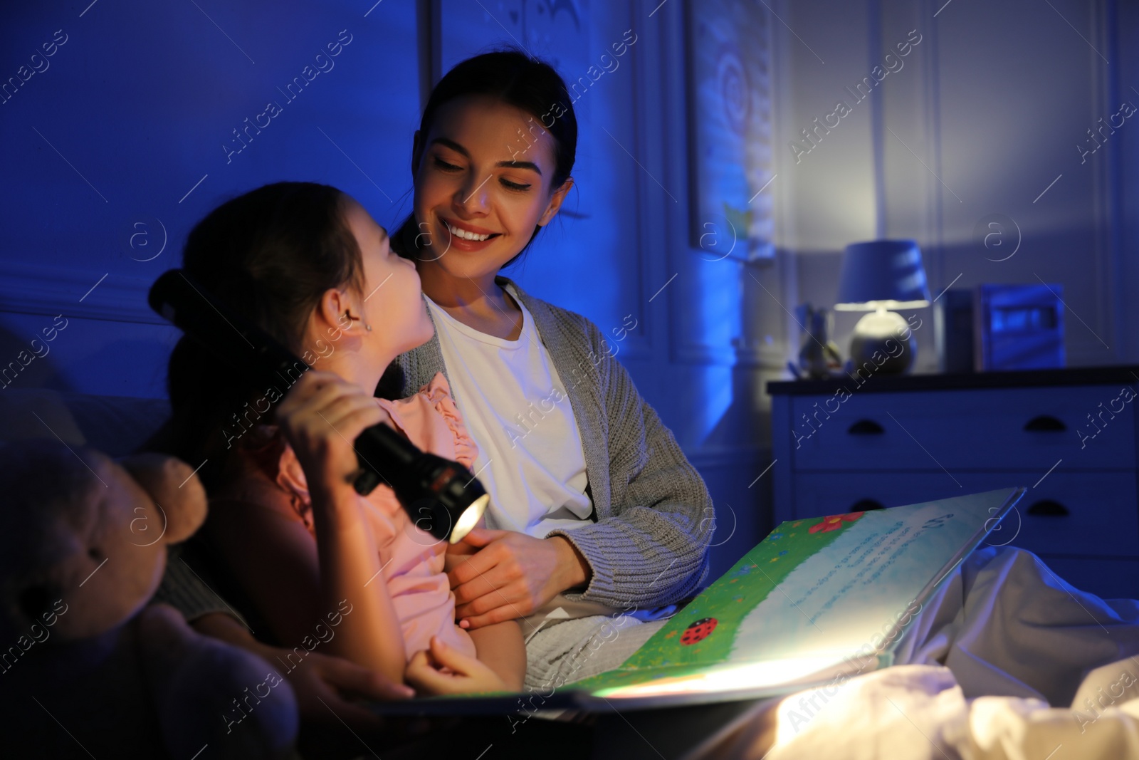 Photo of Mother with little daughter reading fairy tale in dark bedroom