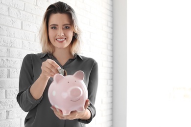 Beautiful woman putting money into piggy bank near brick wall. Space for text