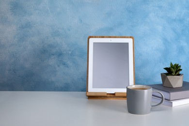 Modern tablet, cup of coffee, books and succulent on white table near light blue wall. Space for design