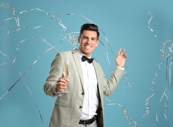 Photo of Happy man with glass of champagne and falling down serpentines on light blue background
