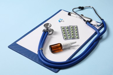 Stethoscope, pills, nasal spray and clipboard on light blue background, closeup. Medical tool