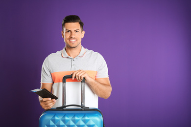 Handsome man with suitcase and ticket in passport for summer trip on purple background. Vacation travel