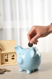 Photo of House model and money on wooden table. Woman putting coin into piggy bank indoors, closeup