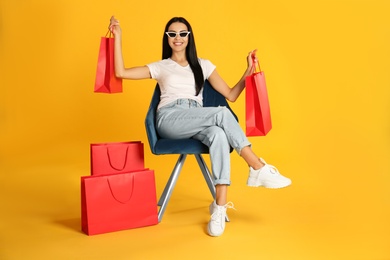 Photo of Beautiful young woman with paper shopping bags in armchair on yellow background
