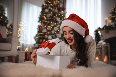 Young woman wearing Santa hat opening Christmas gift on floor at home