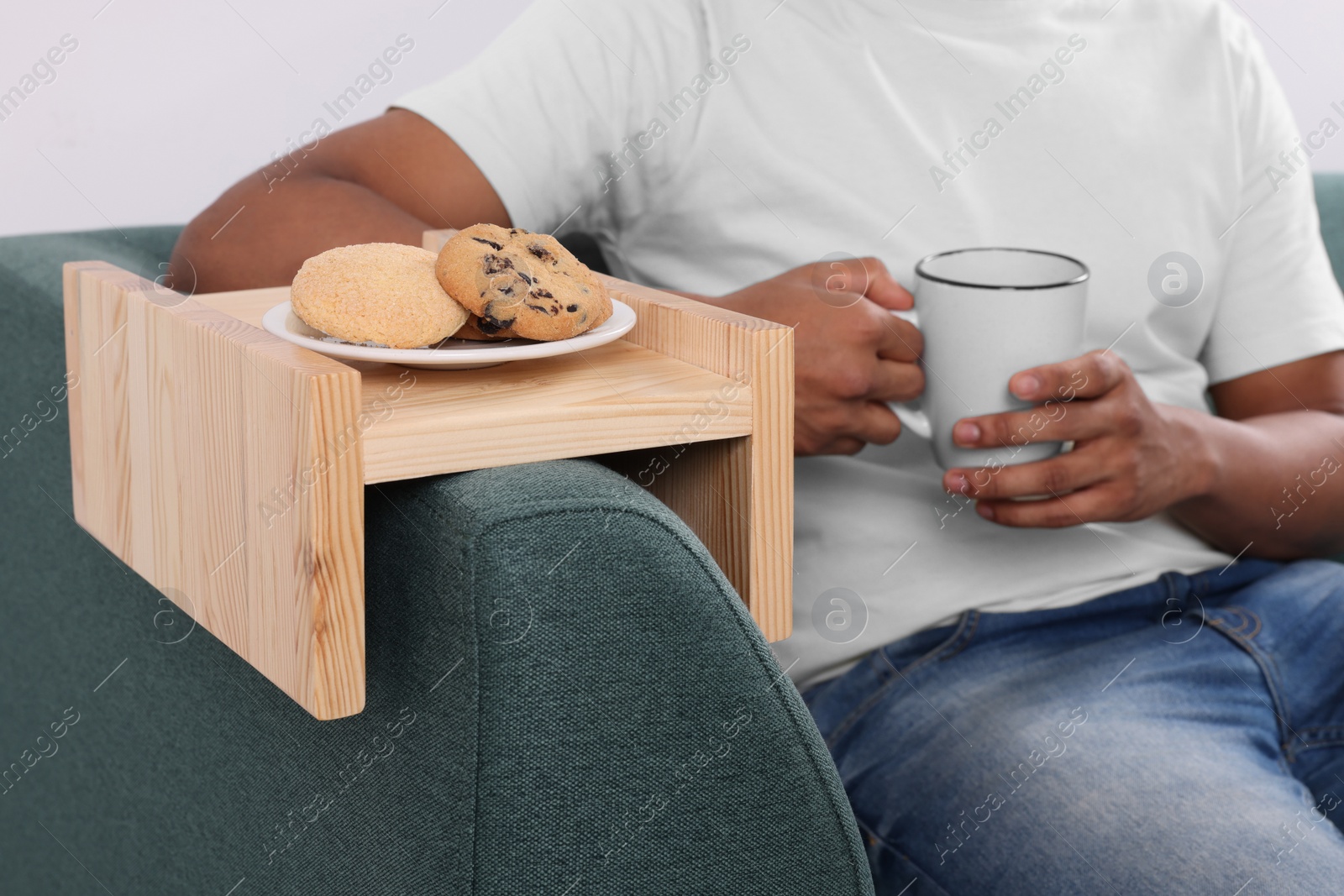 Photo of Cookies on sofa armrest wooden table. Man holding cup of drink at home, closeup