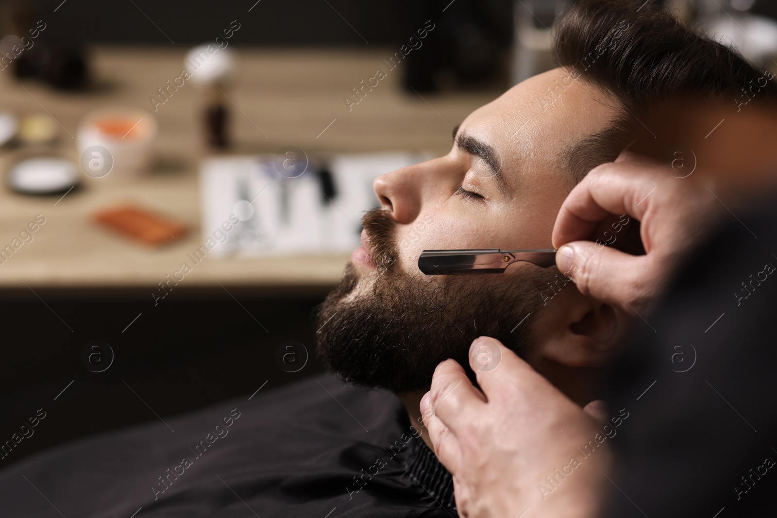 Photo of Professional barber shaving client's beard with blade in barbershop, closeup