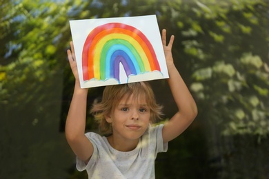 Photo of Little boy with picture of rainbow near window, view from outdoors. Stay at home concept