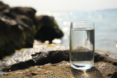 Photo of Glass of fresh water on stone near sea, space for text