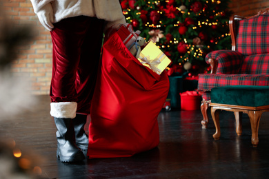Photo of Santa Claus with bag full of Christmas gifts against blurred festive lights, closeup