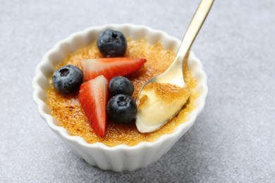 Photo of Delicious creme brulee with berries in bowl and spoon on grey textured table, closeup