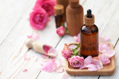 Photo of Rose essential oil and flowers on white wooden table, space for text