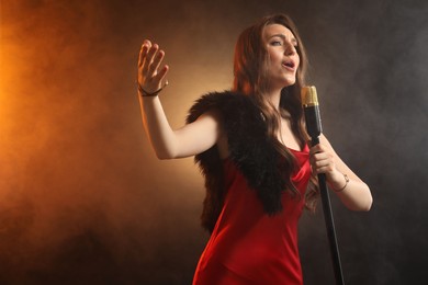 Photo of Beautiful young woman in stylish dress with microphone singing on dark background in color lights and smoke