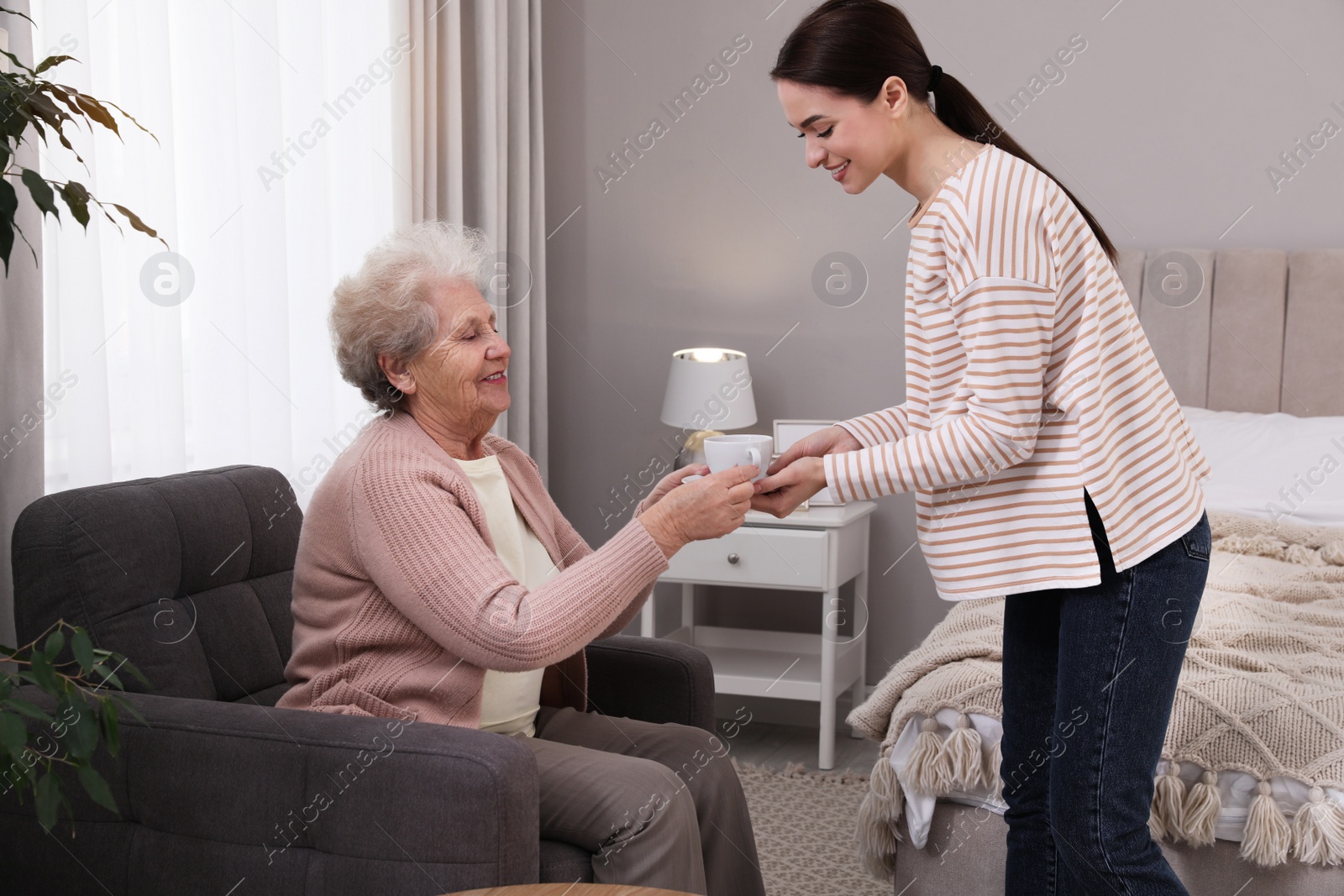 Photo of Young caregiver giving tea to senior woman in room. Home care service