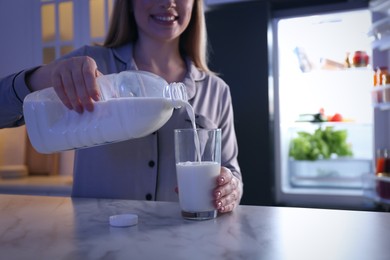 Young woman pouring milk from gallon bottle into glass on white marble table in kitchen at night, closeup