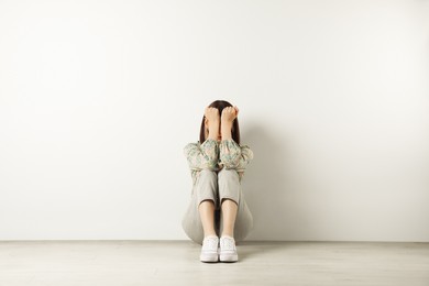 Photo of Young girl hiding face in hands on floor near white wall