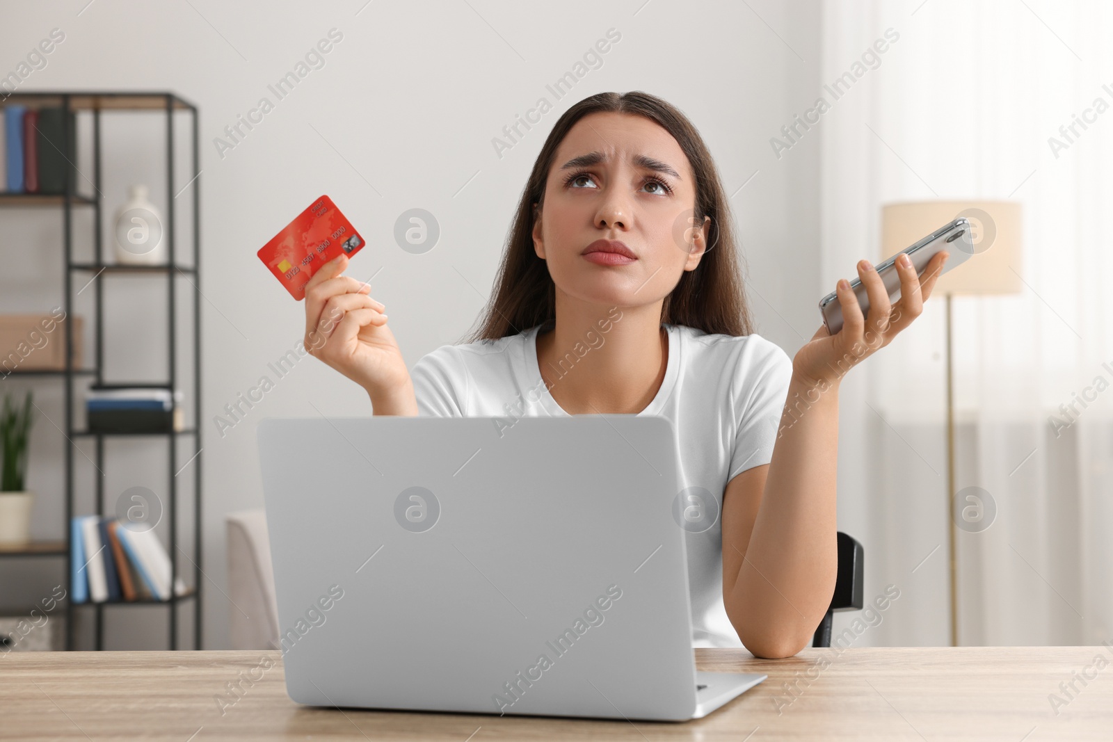 Photo of Confused woman with credit card, smartphone and laptop at table indoors. Be careful - fraud