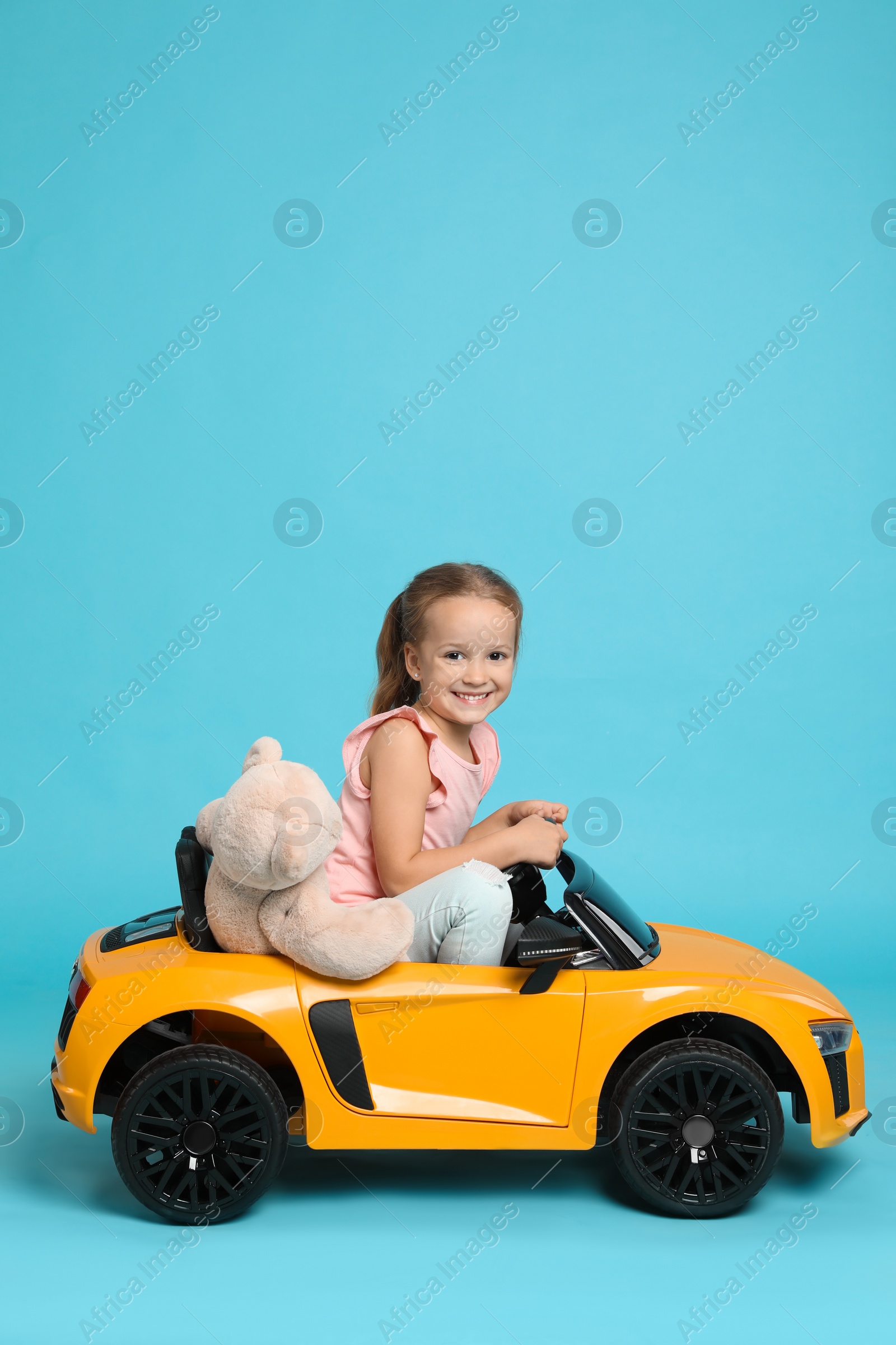 Photo of Cute little girl with toy bear driving children's car on light blue background