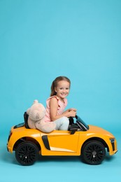 Cute little girl with toy bear driving children's car on light blue background