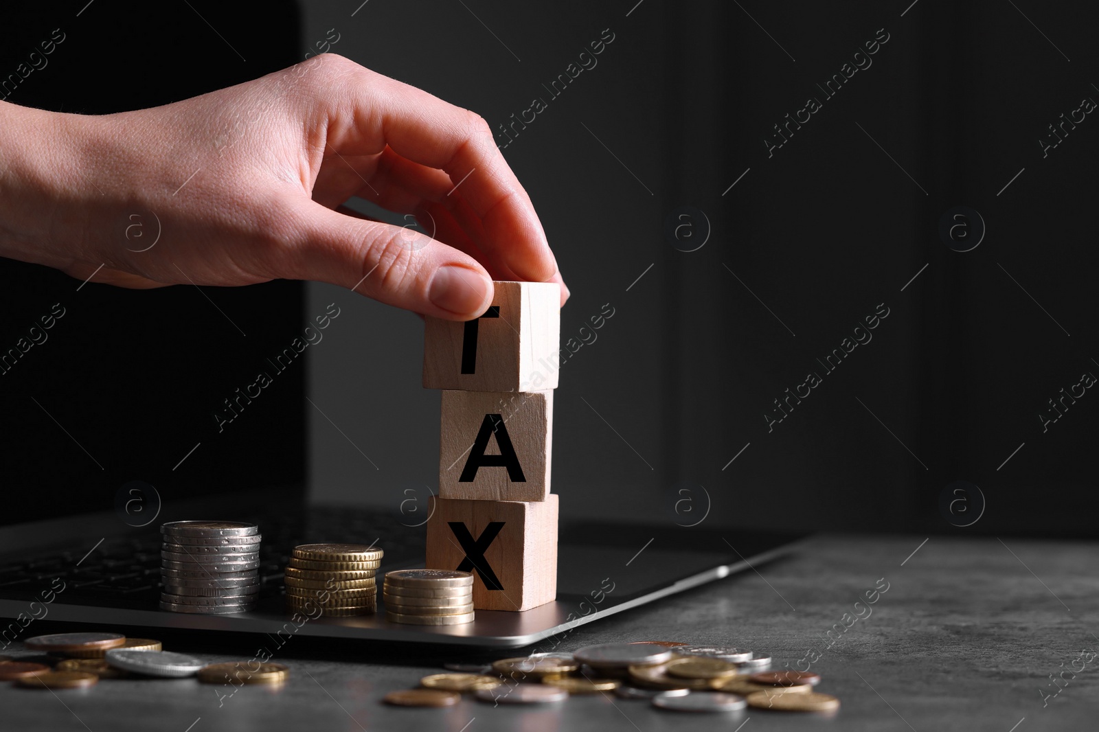 Photo of Woman with word Tax made of wooden cubes, laptop and coins at grey table, closeup. Space for text