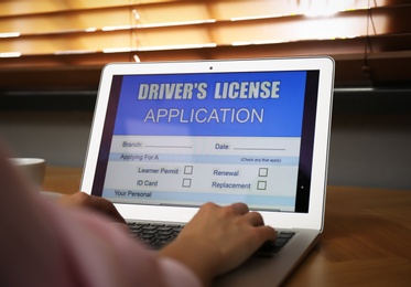 Photo of Woman using laptop to fill driver's license application form at table indoors, closeup