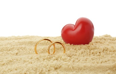 Honeymoon concept. Two golden rings, red wooden heart and sand isolated on white